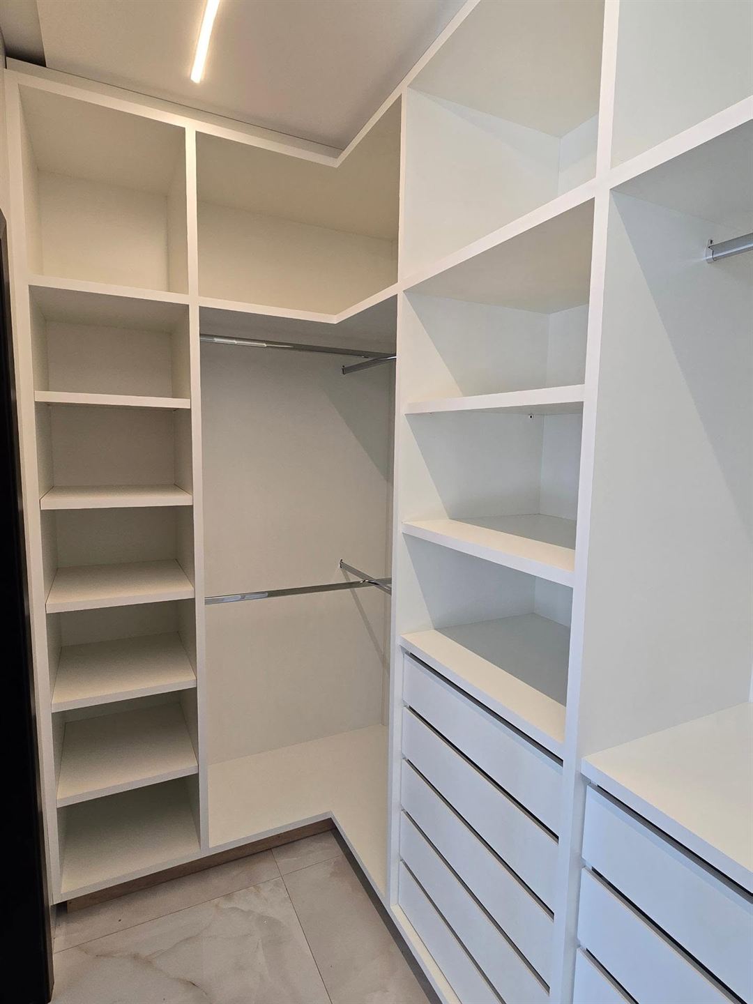 Closet planejado branco em L com prateleiras, cabideiros, gavetas e fita de luz no teto